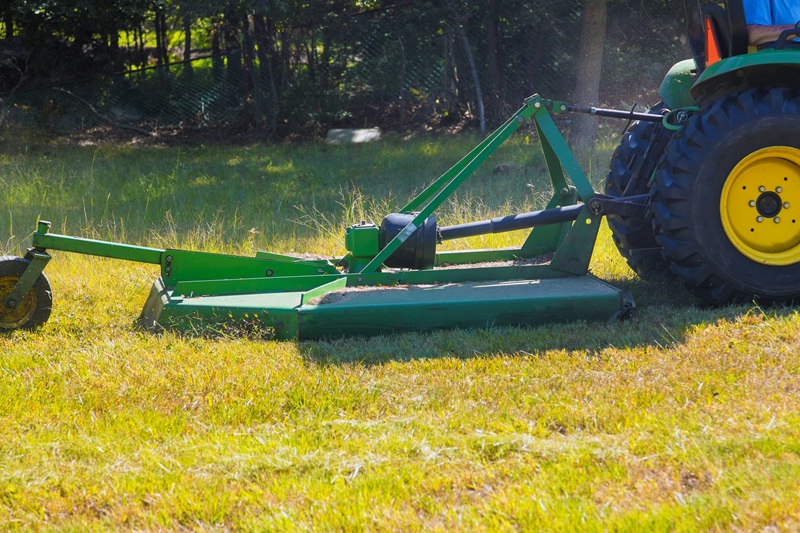 A green tractor with a rotary cutter mowing a slightly overgrown field.